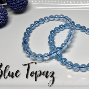May include: Two bracelets made of blue topaz beads are shown on a white plate. The beads are strung on a blue elastic cord. The text "Blue Topaz" is written in a cursive font at the bottom of the image. Decorative blue floral accents are in the background.