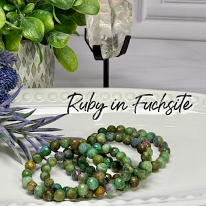 May include: Three beaded bracelets made of green and red gemstones, labeled "Ruby in Fuchsite", rest on a white tray. The background includes a plant, blue flowers, and a crystal.
