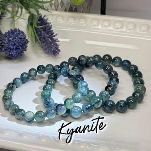 May include: Three bracelets made of blue-green kyanite beads. The beads are round and strung together. The bracelets are displayed on a white tray with decorative edges. The word "Kyanite" is written in cursive.
