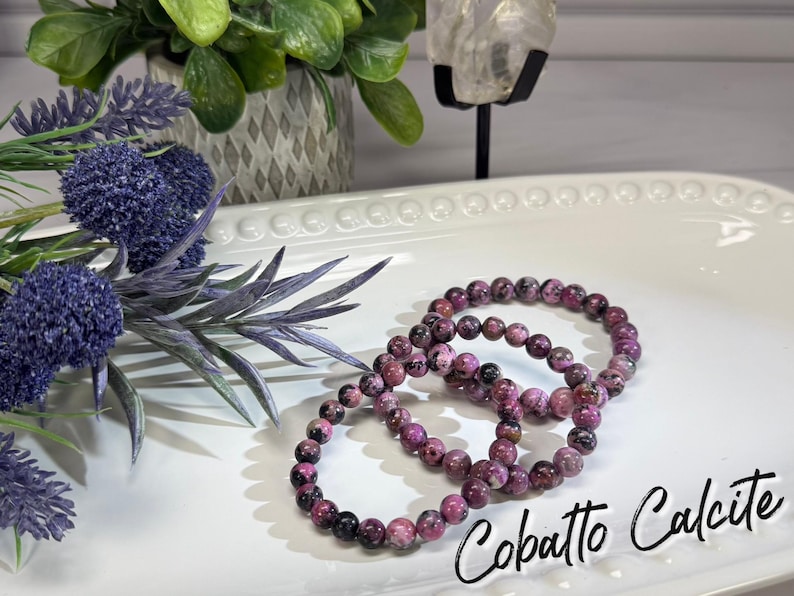 May include: Three bracelets made of round, pink and black beads are displayed on a white, decorative platter. The bracelets are next to purple flowers and greenery. The text "Cobatto Calcite" is written in a script font.
