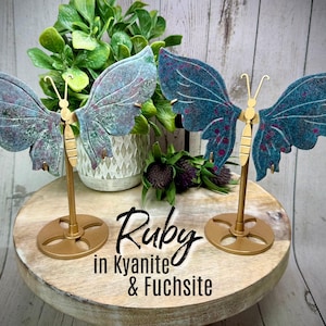 May include: Two butterfly sculptures with teal and purple wings, gold-colored bodies, and round bases. The butterflies are displayed on a wooden stand with the text "Ruby in Kyanite & Fuchsia". Decorative plants are in the background.