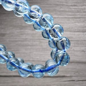May include: A bracelet featuring translucent, light blue, spherical beads threaded on a dark blue cord. The beads have a glassy appearance. The bracelet is displayed against a grey wooden background.
