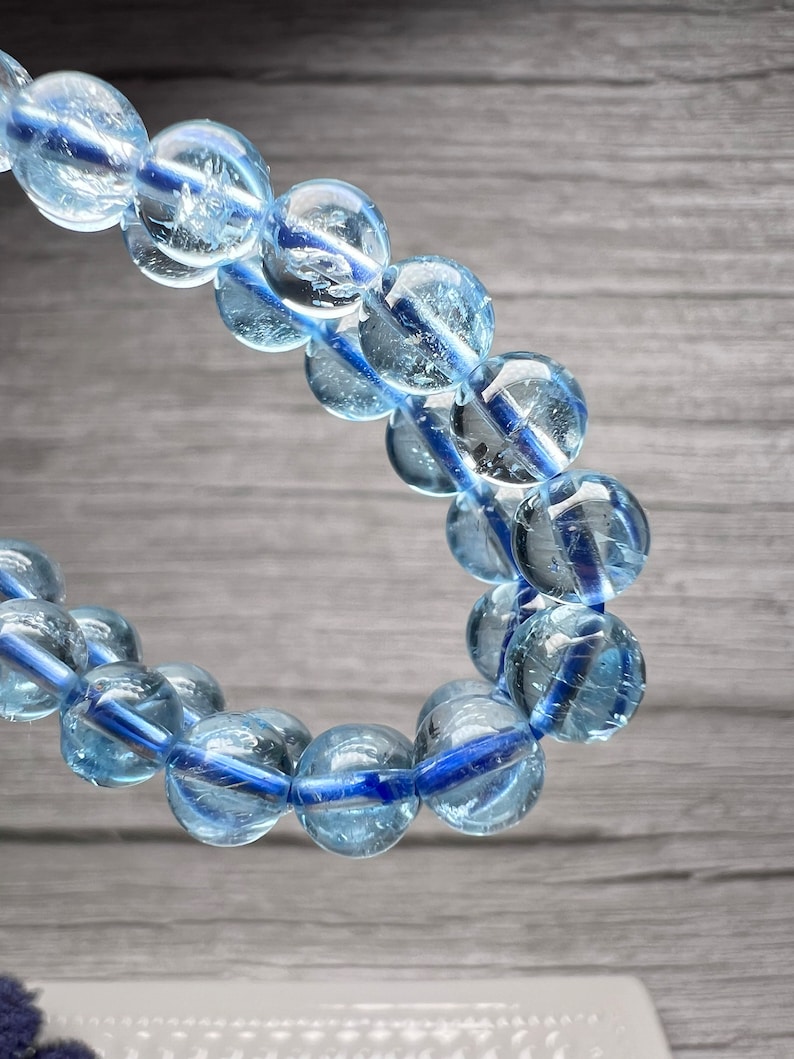 May include: A bracelet featuring translucent, light blue, spherical beads threaded on a dark blue cord. The beads have a glassy appearance. The bracelet is displayed against a grey wooden background.
