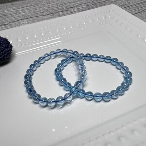May include: Two light blue beaded bracelets. Each bracelet features round, translucent beads strung on a blue elastic cord. The bracelets are displayed on a white, rectangular plate with a decorative edge.