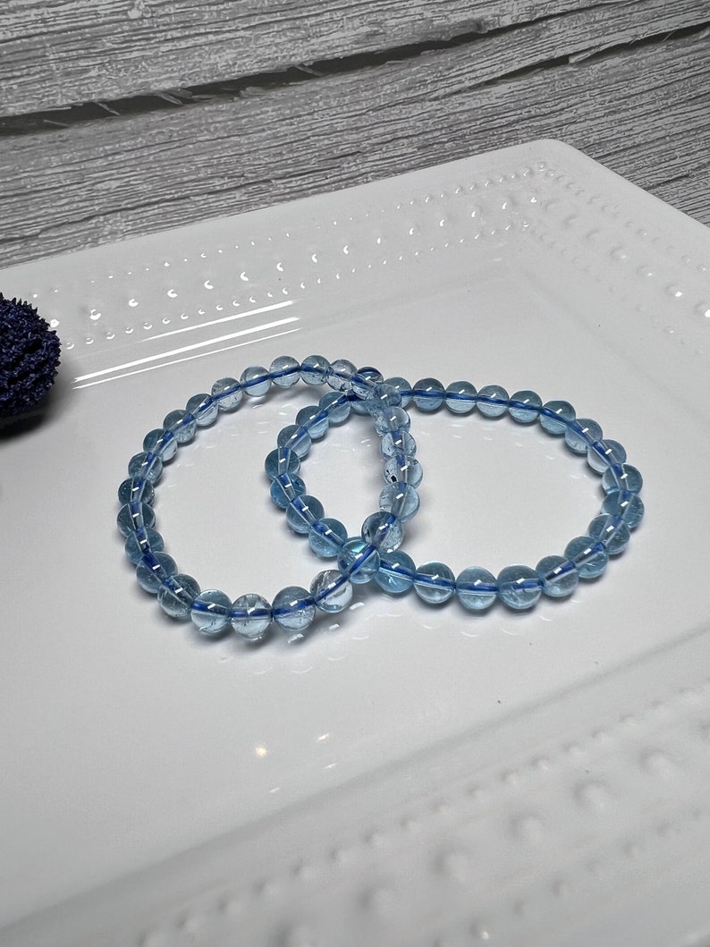 May include: Two light blue beaded bracelets. Each bracelet features round, translucent beads strung on a blue elastic cord. The bracelets are displayed on a white, rectangular plate with a decorative edge.