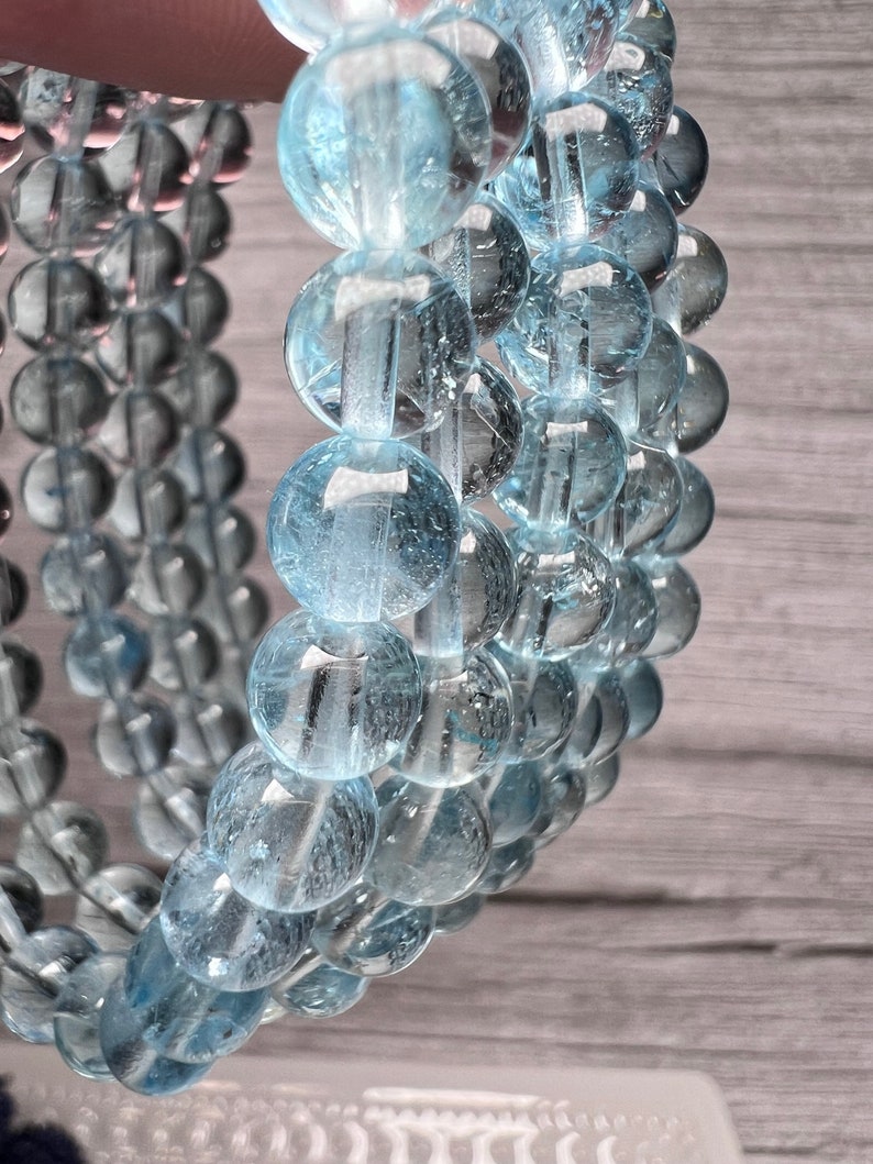 May include: A close-up of a light blue gemstone bracelet with round beads. The beads are translucent and have a slight sparkle.