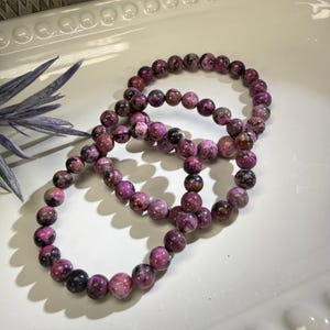 May include: Three beaded bracelets with pink and black stones are arranged on a white surface. The bracelets are made of round, polished beads. A sprig of blue-purple foliage is in the upper left corner.