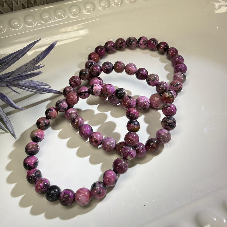 May include: Three beaded bracelets with pink and black stones are arranged on a white surface. The bracelets are made of round, polished beads. A sprig of blue-purple foliage is in the upper left corner.