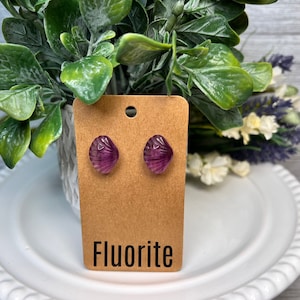 Op de afbeelding: Een paar paarse fluoriet oorbel studs op een bruine kaart met het woord "Fluorite" erop gedrukt.