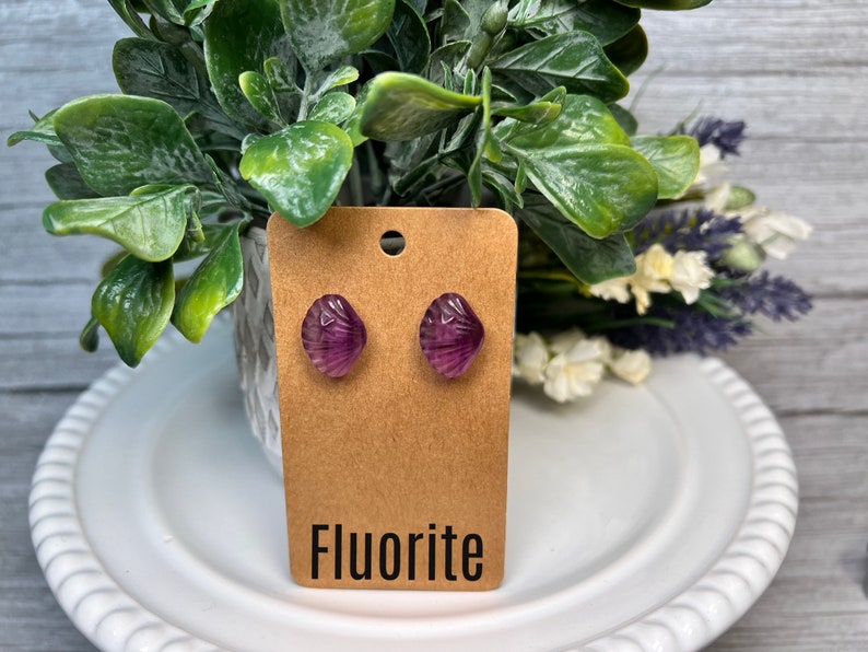 Op de afbeelding: Een paar paarse fluoriet oorbel studs op een bruine kaart met het woord "Fluorite" erop gedrukt.