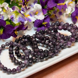 May include: Several dark purple gemstone bracelets with a glossy finish are displayed on a white tray. The bracelets are made of round beads. Purple and white flowers are in the background.