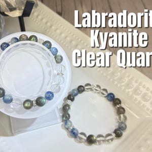 May include: Two beaded bracelets featuring labradorite, kyanite, and clear quartz stones. One bracelet rests on a white display stand, while the other lies on a white, textured surface. The image includes the text "Labradorite Kyanite Clear Quartz."