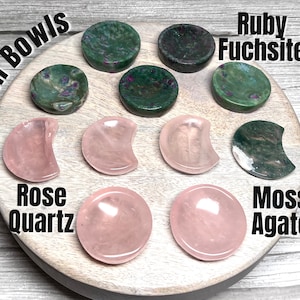 May include: Set of eight mini bowls made of various stones, including rose quartz, moss agate, and ruby fuchsite. The bowls are arranged on a wooden surface. The text "Mini Bowls" is written in the top left corner of the image.