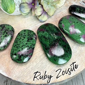 May include: Six polished Ruby Zoisite stones in various sizes, displaying a vibrant green hue speckled with black and patches of pink. The stones are arranged on a wooden surface, with the text "Ruby Zoisite" in a handwritten font.