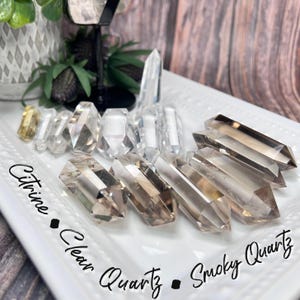 May include: A collection of clear quartz, smoky quartz, and citrine crystal points. The points are arranged on a white surface with the text "Citrine - Clear Quartz - Smoky Quartz" written below them.