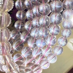 May include: Close-up of a strand of clear, round beads with a pinkish-purple string running through them. The beads have a reflective, crystalline appearance, and the string is visible within each bead. The beads are arranged in a neat, parallel pattern.