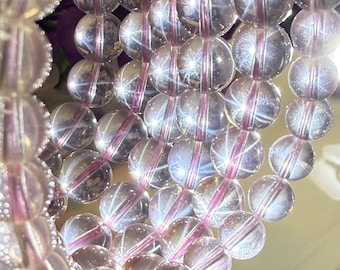 Può includere: Primo piano di una collana di perline rotonde e trasparenti con un filo rosa-viola che le attraversa. Le perline hanno un aspetto riflettente e cristallino, e il filo è visibile all'interno di ogni perlina. Le perline sono disposte in un motivo ordinato e parallelo.