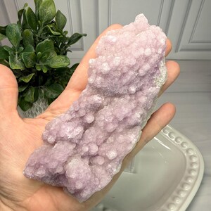 May include: A large, light purple crystal cluster with a bubbly texture. The crystal is held in a hand, showcasing its size and color. The crystal's surface has a unique, organic appearance, with a mix of smooth and textured areas.