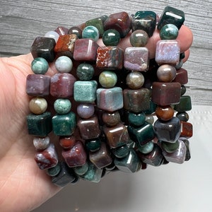 May include: A stack of bracelets made with green, brown, and red jasper beads. The beads are all square shaped and have a polished finish.