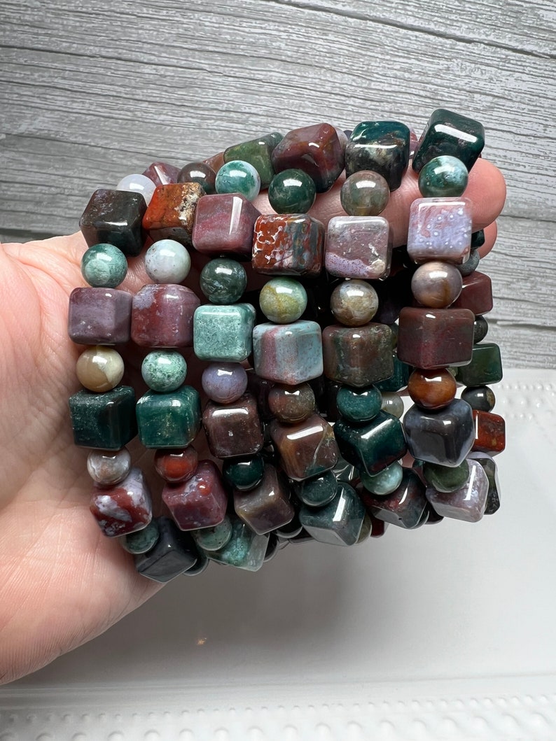 May include: A stack of bracelets made with green, brown, and red jasper beads. The beads are all square shaped and have a polished finish.