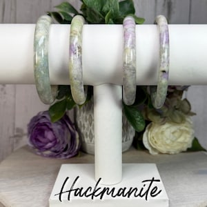 May include: Four Hackmanite bracelets displayed on a white stand. The bracelets are a mix of pale green and lavender hues, with a marbled appearance. The stand has the word "Hackmanite" in a script font.