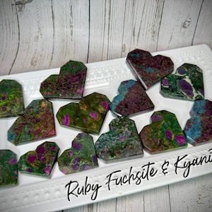 May include: A collection of heart-shaped stones in shades of green, purple, and blue, arranged on a white rectangular plate. The stones are labeled "Ruby Fuchsite & Kyanite". The background is a light wood grain.
