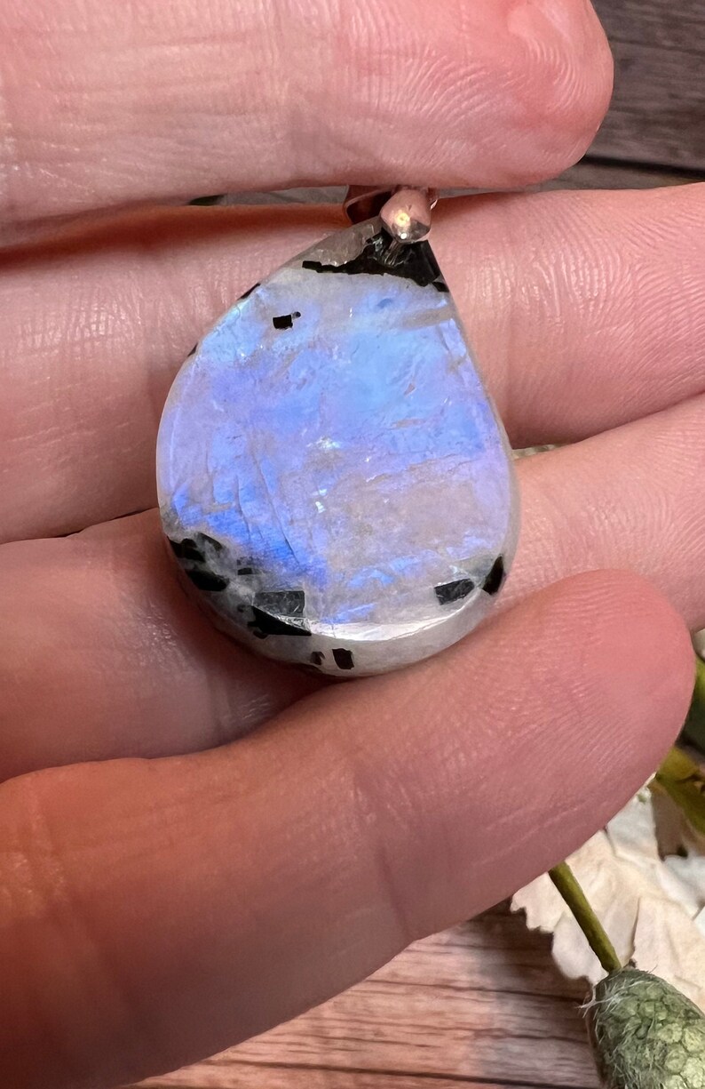 May include: A teardrop-shaped iridescent moonstone pendant with black inclusions. The pendant is set in silver and has a polished finish.