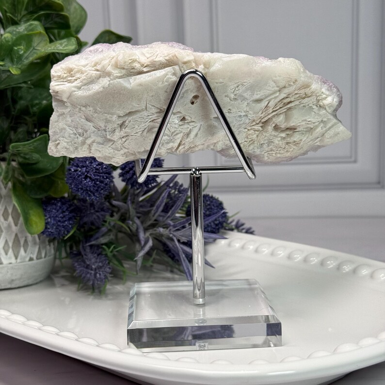 May include: A light-colored, textured stone displayed on a silver metal stand with a clear acrylic base. The stone is held by a triangular support. The background includes greenery and purple flowers, all set on a white tray.