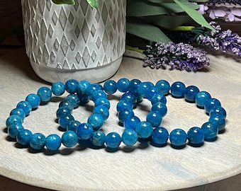 May include: Three blue apatite beaded bracelets stacked on a wooden surface. The beads are round and have a light blue color with a slight shimmer.