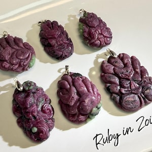 May include: Six carved ruby in zoisite pendants with silver-colored bails. The pendants are a deep burgundy color with green accents. The pendants are carved with a detailed, organic design. The text "Ruby in Zoisite" is written in black script.
