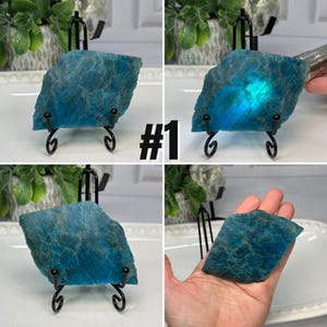 May include: A flat, diamond-shaped piece of blue apatite mineral with natural textured surfaces and mottled dark inclusions. The stone is displayed on a black metal wire stand and also shown held in a palm, with one view illuminated by light.