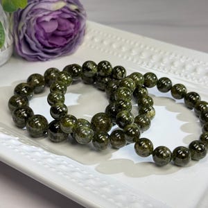 May include: Two elastic bracelets made of polished, spherical dark olive green beads with subtle mottled patterns. The beads have a glossy finish and are strung together in a circular formation, showcasing deep forest green tones and natural variations.