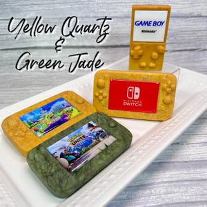 May include: Three miniature handheld game consoles made of yellow quartz and green jade. The consoles are decorated with images of video game characters and logos. The consoles are labeled "GAME BOY", "NINTENDO SWITCH", and "Pokémon UNITE".