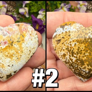 May include: Two heart-shaped stones with unique patterns. One features a mix of white, yellow, and pink hues, while the other displays a blend of yellow and brown. The stones are held in a hand, showcasing their natural beauty.