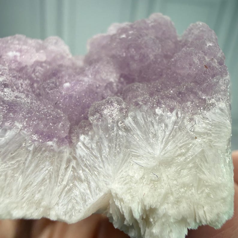 May include: Close-up of a natural mineral specimen. The top portion is a soft purple hue, transitioning to a white, crystalline base. The texture is rough and uneven, with visible crystal formations.