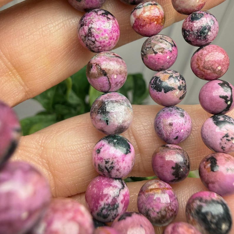 May include: Close-up of a strand of round gemstone beads. The beads are primarily pink with dark gray and brown inclusions. The beads are approximately 0.3 inches in diameter. The beads are strung together.
