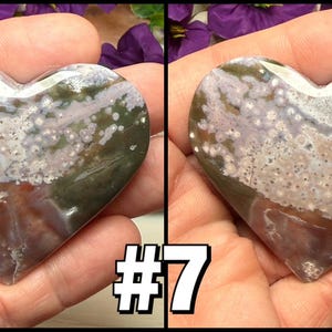 May include: A heart-shaped stone with a polished surface, featuring a mix of colors including white, green, brown, and purple. The stone is held in a hand, with the number "7" in a white outlined font in the lower right corner.