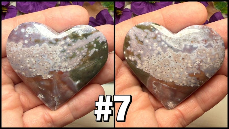 May include: A heart-shaped stone with a polished surface, featuring a mix of colors including white, green, brown, and purple. The stone is held in a hand, with the number "7" in a white outlined font in the lower right corner.
