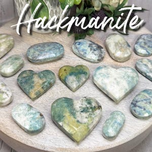 May include: A collection of hackmanite stones in various shapes, including ovals and hearts, arranged on a wooden surface. The stones display a mix of colors, including white, blue, and green, with the word "Hackmanite" in white script at the top.