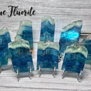 May include: Eight blue fluorite crystal specimens on metal stands. The crystals are translucent and have a layered appearance. The text "Blue Fluorite" is written in black cursive at the top of the image.