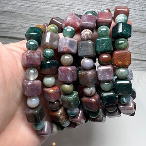 May include: A stack of bracelets made with multi-colored agate beads. The beads are a mix of round and square shapes. The bracelets are a mix of green, brown, red, and purple.
