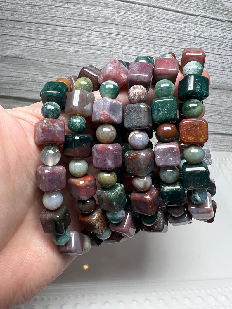 May include: A stack of bracelets made with multi-colored agate beads. The beads are a mix of round and square shapes. The bracelets are a mix of green, brown, red, and purple.