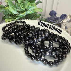 May include: A collection of elastic bracelets made from polished black hypersthene beads. The beads feature a subtle metallic sheen and are arranged in circular strands. The text "Hypersthene" is printed in black on the white surface.