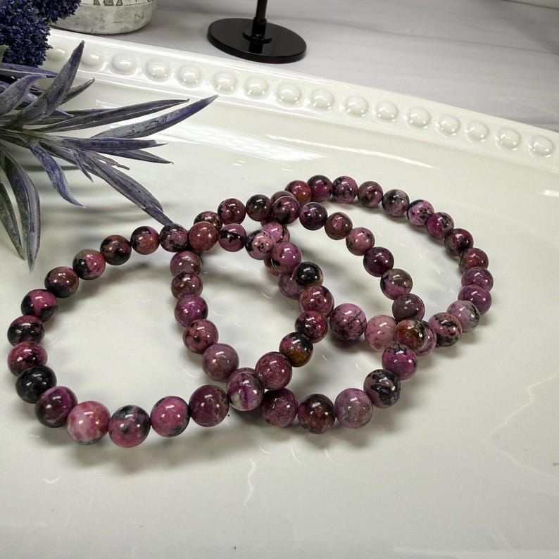 May include: Three beaded bracelets with a mix of dark pink, black, and white round beads. The bracelets are displayed on a white, oval-shaped tray. The beads have a marbled appearance.