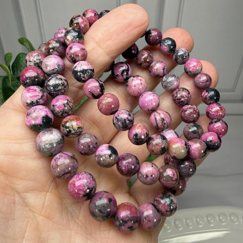May include: A collection of pink and black gemstone bead bracelets. The beads are round and have a marbled appearance, with varying shades of pink and black. The bracelets are held in a hand, showcasing the natural stone's colors and textures.