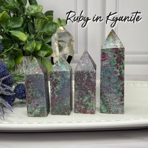 May include: Four Ruby in Kyanite crystal towers. The crystals are a mix of deep red, green, and gray. The crystals are displayed on a white tray. The text "Ruby in Kyanite" is written in cursive at the top of the image.