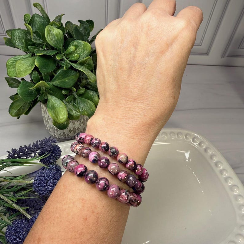 May include: Three bracelets with pink and black beads are stacked on a wrist. The beads have a marbled appearance. The background includes a green plant in a gray pot, purple flowers, and a white decorative plate.