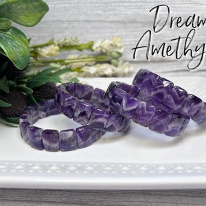 May include: Three amethyst gemstone bracelets with a chevron pattern. The bracelets are stacked on a white plate. The text "Dream Amethyst" is written in white on a gray background.