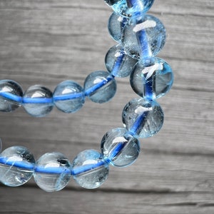 May include: A close-up of a light blue beaded bracelet. The round, transparent beads have a blue stripe. The bracelet is strung on a blue cord. The background is a grey wooden surface. The beads are approximately 1 cm in diameter.