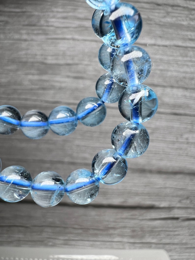 May include: A close-up of a light blue beaded bracelet. The round, transparent beads have a blue stripe. The bracelet is strung on a blue cord. The background is a grey wooden surface. The beads are approximately 1 cm in diameter.